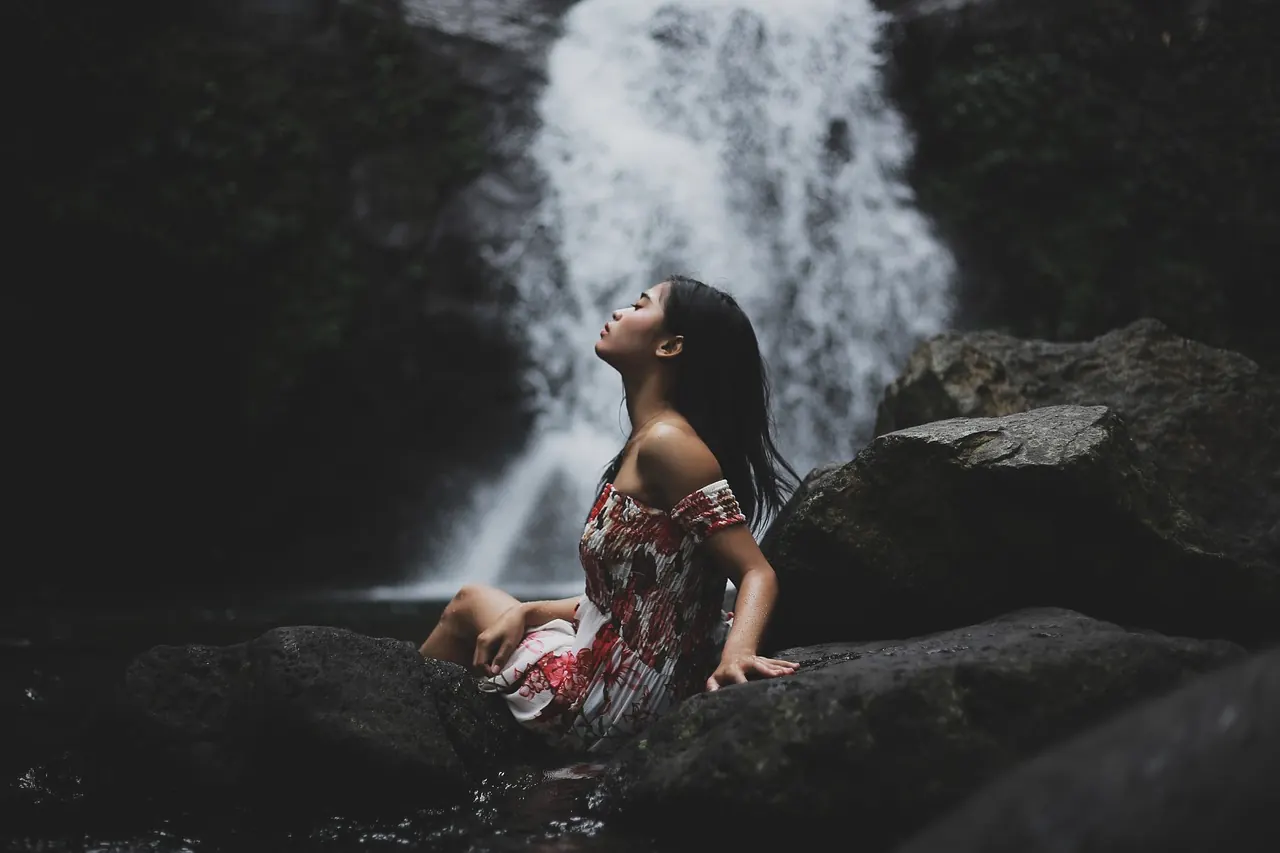 Frau sitzt entspannt auf einem Felsen vor einem Wasserfall, den Kopf nach oben gewandt